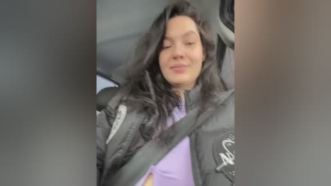 Video of a woman with long, dark hair, wearing a grey puffer jacket and purple shirt, seated in a car with a seatbelt across her chest, smiling slightly.