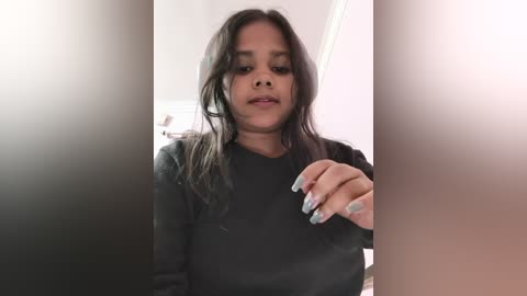 Video of a young South Asian girl with long dark hair, wearing a black sweater, smiling. The background is blurry, featuring a white wall and a door.