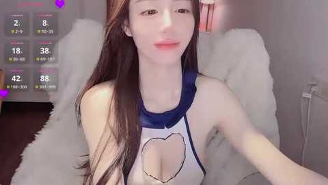 A video of an Asian woman with long brown hair, fair skin, and a slender figure, wearing a revealing, white-and-blue sports bra with a heart cutout, sitting on a fluffy white blanket.