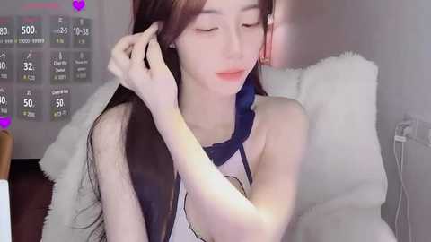 A video of a young East Asian woman with long brown hair, fair skin, and delicate features, sitting on a white couch. She wears a sleeveless top and is adjusting her hair.