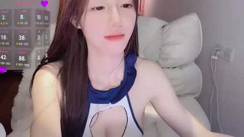 Video of an East Asian woman with long, straight black hair, fair skin, and a slender physique. She wears a revealing, white-and-blue top with a large cutout, seated on a white leather couch. The background shows a wall-mounted TV displaying a calendar.