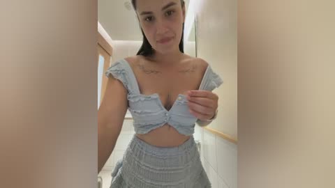 A video of a young woman with light skin, straight black hair, and a slender build, wearing a light blue ruffled crop top and matching skirt, standing in a hallway with cream-colored walls.