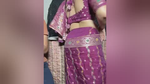 Video of a woman in a vibrant purple saree with gold embroidery, standing beside a person in a black shirt, indoors, blurred background.
