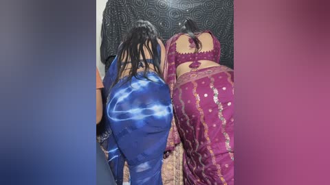 Video of two women in colorful, patterned sarees, one in blue, the other in maroon, kneeling in front of a black, intricately designed wall.