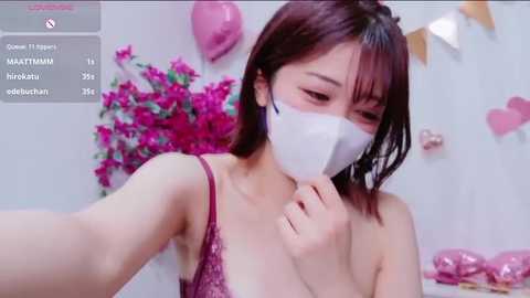 A video of an East Asian woman with straight, dark brown hair, wearing a white mask and a pink patterned spaghetti-strap top. Background includes pink balloons, heart banners, and a pink flower arrangement.