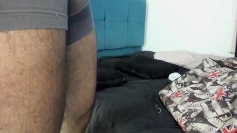 Video of a close-up view of a man's hairy leg in grey shorts, standing beside a bed with a black comforter and a Union Jack-patterned pillow. Background shows a turquoise padded headboard and white walls.