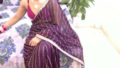 Video of a woman in a purple sari with white stripes, sitting on a bed with blue and white floral sheets, wearing a red bra and red bangles.