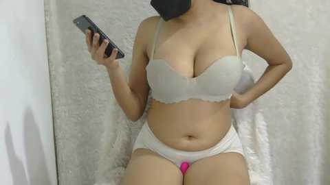 A video of a curvy, light-skinned woman with medium-sized breasts in white lingerie, holding a phone. She stands in front of a white, textured curtain background.