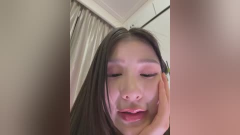 A close-up video of an Asian woman with long, straight black hair, light skin, and closed eyes, leaning her head on her hand. The background features beige curtains and white walls.