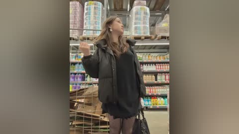 Video of an Asian woman with long hair in a grocery store, wearing a black oversized coat and holding a black handbag, standing near stacked toilet paper rolls and colorful bottles.