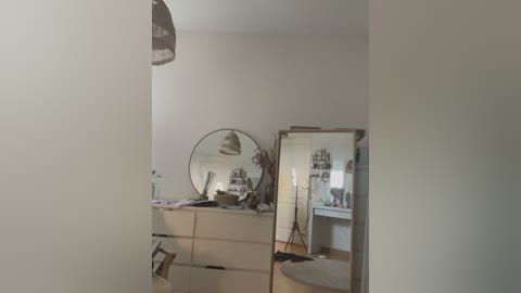 Video of a minimalist bathroom with a white dresser, round mirror, silver-framed door, and a hanging pendant light. The room is bright with a neutral color palette, creating a clean and modern atmosphere.