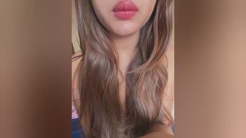 Close-up video of a woman with long, wavy brown hair and full, pink lips, wearing a pink top. Background is blurred and neutral-toned.