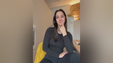 Video of a young woman with long dark hair, fair skin, and slender physique, wearing a black long-sleeve top, seated in a modern kitchen with beige walls and a wooden ceiling.