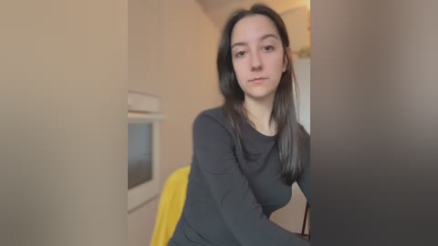 A video of a young Caucasian woman with long, straight black hair, wearing a dark gray long-sleeve shirt, sitting indoors. The background features beige walls, a microwave, and a yellow towel.