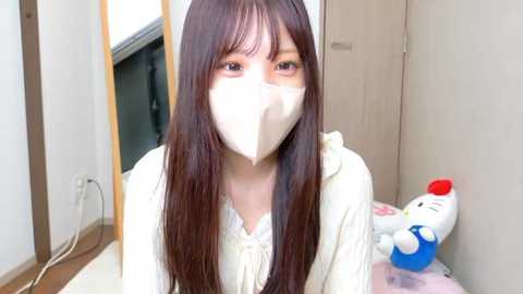 A video of an Asian woman with long brown hair, wearing a white face mask and a cream-colored top, sitting in a light-colored room with a wooden floor and a plush toy on a table.