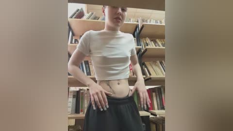 Video of a slender, light-skinned woman with short, dark hair, wearing a white crop top and black pants, standing in a library with bookshelves in the background.
