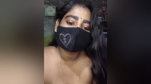 Video of a South Asian woman with long black hair, wearing a black face mask with a heart design, and a thin gold necklace. Background shows a wall clock, a green couch, and a cluttered bookshelf.