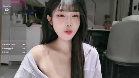 A video of an East Asian woman with long black hair and pale skin, wearing a white off-shoulder blouse, in a modern office setting.