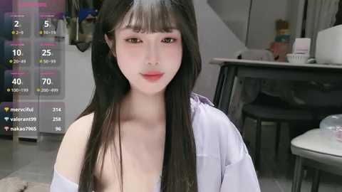 Video of a young Asian woman with long black hair, pale skin, and pink lips, wearing a white shirt, sitting in a modern kitchen.