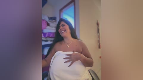 Media: Video of a smiling, pregnant Latina woman with long black hair, wearing a white towel, in a home with stacked laundry and a window.