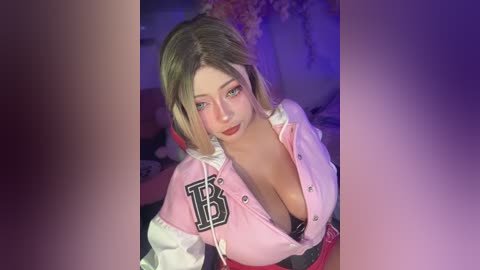 Video of a light-skinned woman with blonde hair, wearing a low-cut pink jacket with a black \"B\" logo, posing seductively in a dimly lit bedroom.