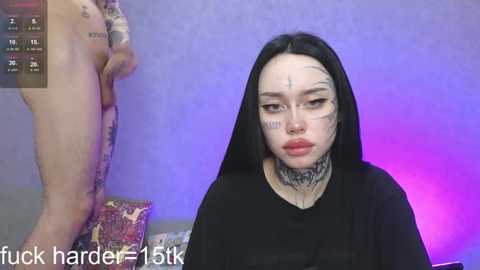 Media: Video of a pale-skinned, black-haired woman with heavy makeup, including black ink tattoos on her face, sitting in a dimly lit room with a purple light. She wears a black top, and a nude figure stands behind her.