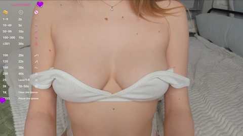 A video of a light-skinned woman with auburn hair, wearing a white, off-shoulder crop top that reveals ample cleavage, standing in a bedroom with a white bed and green pillow.