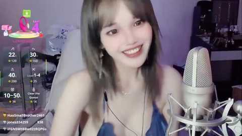 Video of a smiling young woman with shoulder-length gray hair, wearing a blue halter top, seated in front of a microphone on a dimly-lit desk, surrounded by colorful digital overlays.