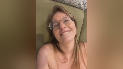 A close-up video of a smiling, topless Caucasian woman with light skin and shoulder-length brown hair, wearing glasses, lying on a green leather couch.