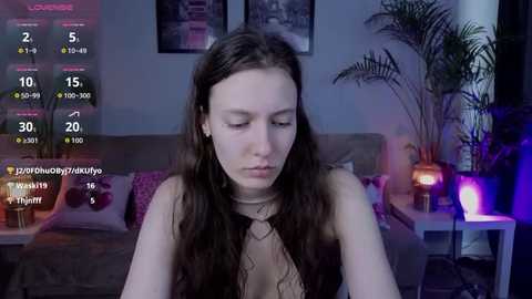 Media: Video of a young woman with long brown hair, wearing a black lace top, sitting indoors with a dimly lit room in the background.