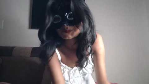Media: A video of a woman with long black hair, wearing a black lace mask and a white tank top, sitting on a couch in a dimly lit room.