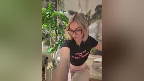 Video of a young woman with short blonde hair, wearing black-rimmed glasses, a black crop top with pink text, and pink shorts, seated in a room with green plants and a patterned wall.