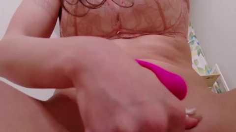 A video of a nude woman with fair skin and red hair, using a bright pink vibrator on her vulva, in a bathroom with floral wallpaper.