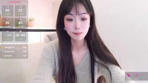 Video of an East Asian woman with long black hair, fair skin, and a slim physique, wearing a light grey sweater. Background includes a modern room with a light-colored wall and a potted plant.