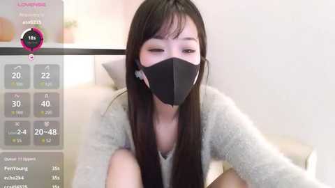 A young Asian woman with long black hair and a black face mask, wearing a grey sweater, sits indoors, with a digital thermometer displaying her temperature on the left.