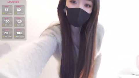 A video of an East Asian woman with long brown hair, wearing a black face mask and a gray coat, taking a selfie indoors.