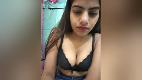 Video of a young woman with medium skin tone, wearing a black lace bra, her long brown hair cascading over her shoulders, in a bathroom with a tiled wall and a pink towel in the background.