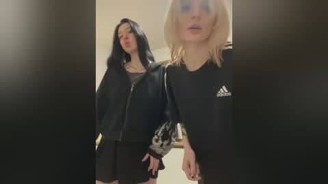 Video of two young women with pale skin and shoulder-length hair, one with black, the other blonde. They wear black Adidas jackets, standing in a dimly lit room with beige walls.