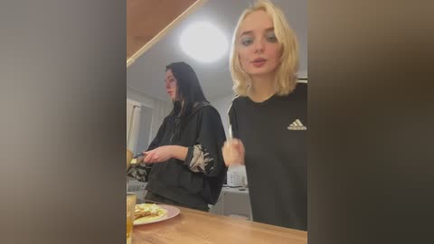 Video of a blonde woman in a black Adidas sweatshirt, looking surprised, standing next to a woman with long black hair in a black robe, both at a kitchen counter.