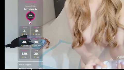 Video of a topless woman with long, wavy blonde hair, wearing a black bra, standing in a bathroom with a white bathtub and blue tiles. A digital overlay shows her \"Envy Score\" of 26%.