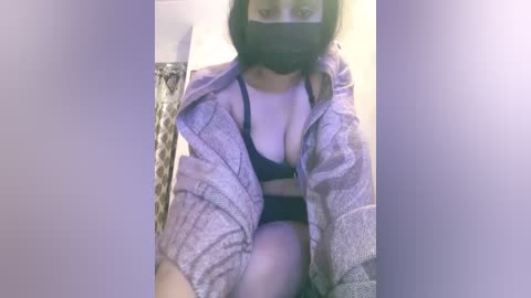 A video of a woman in a black bra and gray cardigan, wearing a black face mask, sitting indoors.