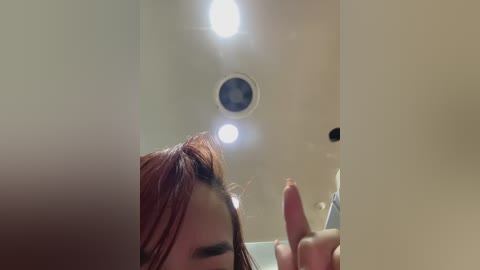 A video of a woman with wet, red hair, partially seen, holding a pink dildo, taken from a low angle in a dimly lit room with a ceiling light.