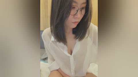 Media: A video of an Asian woman with straight black hair and glasses, wearing a sheer white blouse that reveals her cleavage, seated indoors.