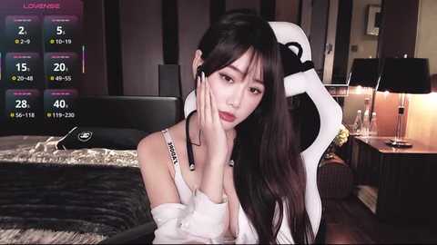 Video of a young East Asian woman with long black hair, wearing a white off-shoulder top, sitting on a white gaming chair in a dimly lit room with a bed, lamps, and a digital clock.