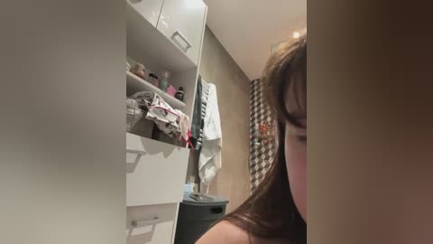 A video shows a woman with long brown hair, partially cropped, standing in a modern, organized bathroom. White cabinets with drawers hold toiletries, towels, and a black trash bin. The background includes beige tiles and a towel rack with hanging towels.
