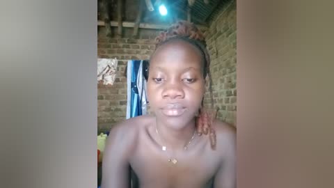 A video of a topless young Black woman with light skin, pink dreadlocks, and a necklace, standing in a rustic, dimly lit room with brick walls and a wooden ceiling.