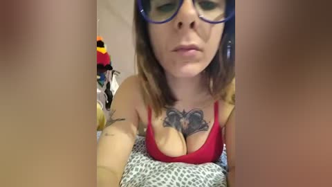 Video of a light-skinned woman with glasses, medium-length brown hair, wearing a red bra revealing cleavage and a leopard-print top. Background includes a colorful stuffed animal and clothing.