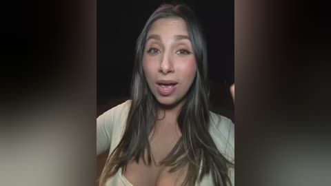 Video of a woman with long, dark hair, fair skin, and a medium build. She has a surprised expression and is wearing a beige top. The background is dark and blurry.