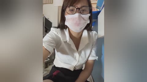 Video of an East Asian woman with straight brown hair, wearing a white blouse, black skirt, and a surgical mask, seated in a room with blue and white walls, a desk, and medical equipment.
