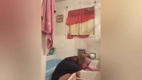 Video of a young person with long brown hair, wearing a black shirt, leaning over a bathtub with a blue tub insert, in a small, cluttered bathroom with a pink towel hanging and a red polka-dotted curtain.